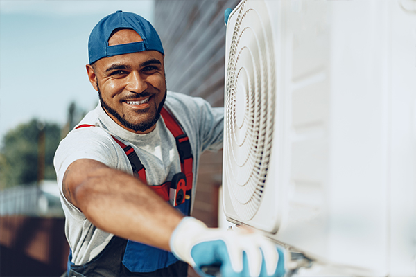 A man is fixing a commercial HVAC unit and smiles at the camera. He is excited to have started his own HVAC business. 